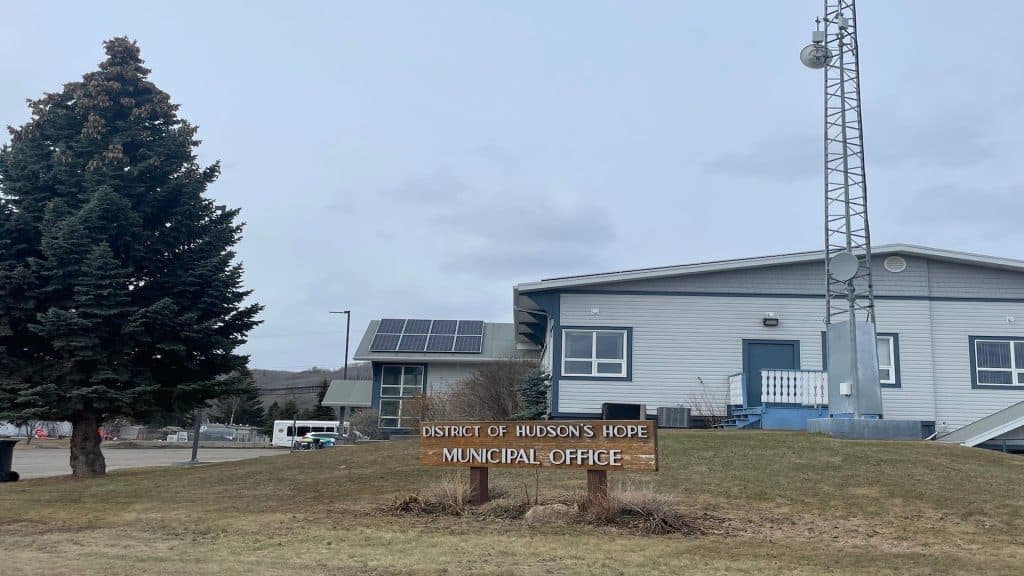 A picture of a building with green trim next to a large evergreen tree with a wooden sign out front that reads "District of Hudson's Hope Municipal Office"