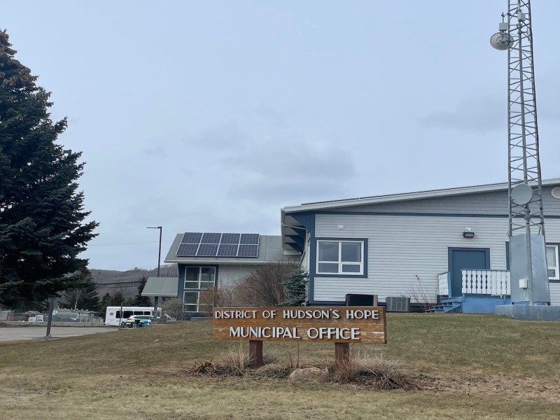 A picture of a building with green trim next to a large evergreen tree with a wooden sign out front that reads "District of Hudson's Hope Municipal Office"