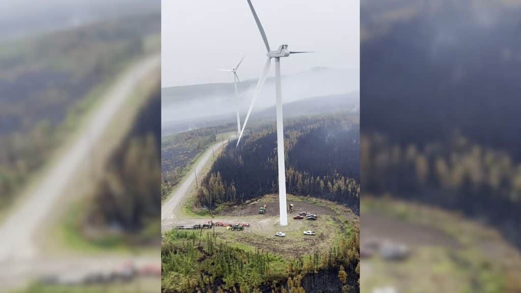 An aerial shot of a white wind turbine surrounded by vehicles, a forest, and wildfire smoke.