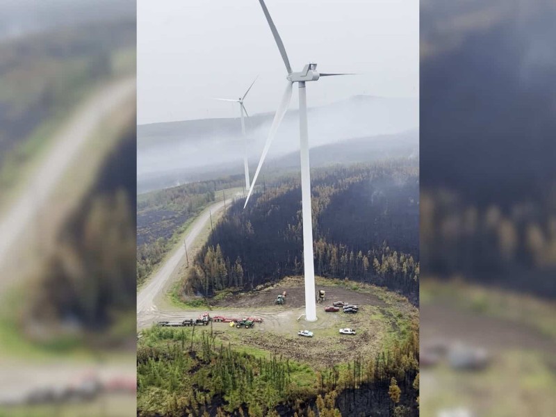 An aerial shot of a white wind turbine surrounded by vehicles, a forest, and wildfire smoke.