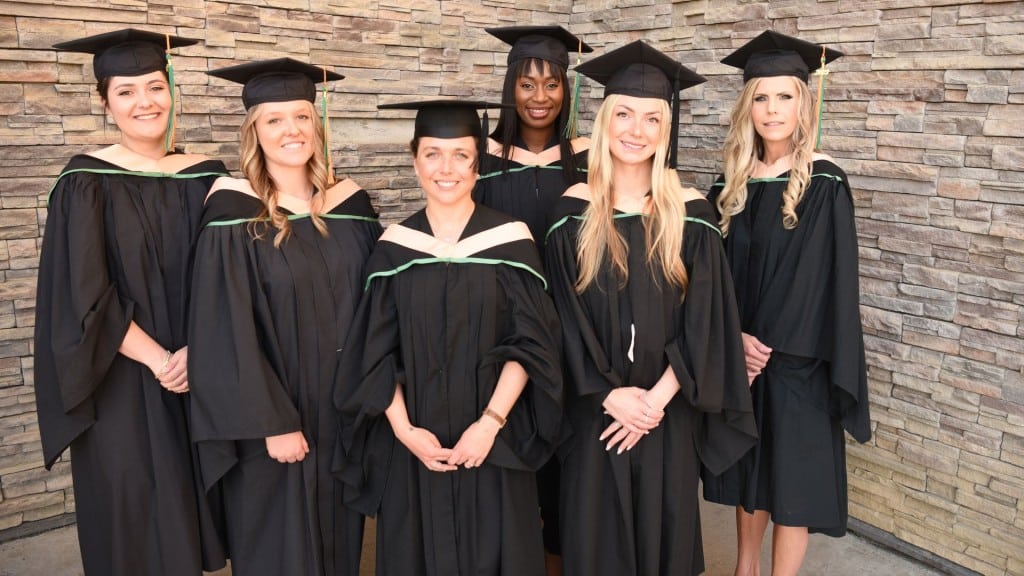 A group of six women standing in graduation gowns in front of a brick building.
