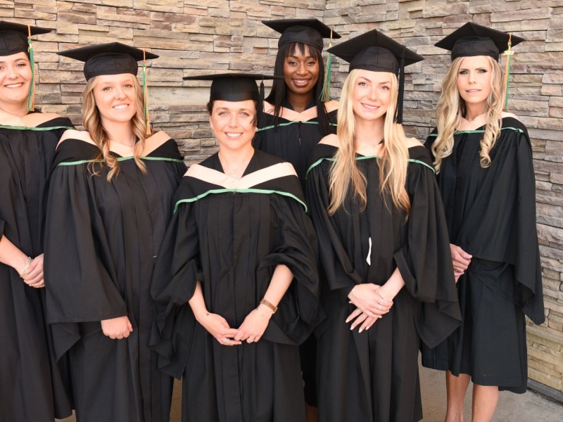 A group of six women standing in graduation gowns in front of a brick building.