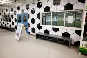 A picture of a hallway with soccer ball wallpaper, a grey bench and windows that look onto an indoor soccer field.