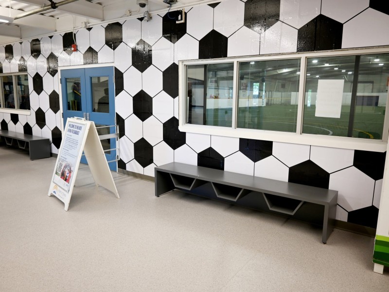 A picture of a hallway with soccer ball wallpaper, a grey bench and windows that look onto an indoor soccer field.
