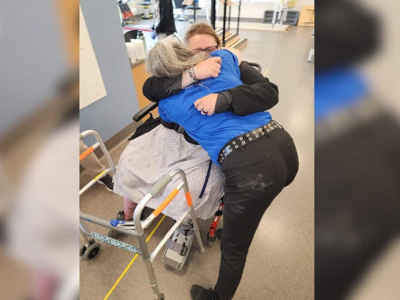 A woman in a wheelchair wearing a cast hugs an older woman who is standing over her.