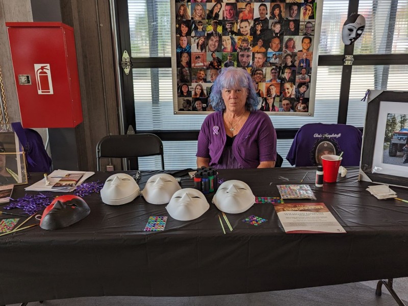 A woman with purple hair sits at a table with a photo of her deceased son and his friend at an overdose awareness event.