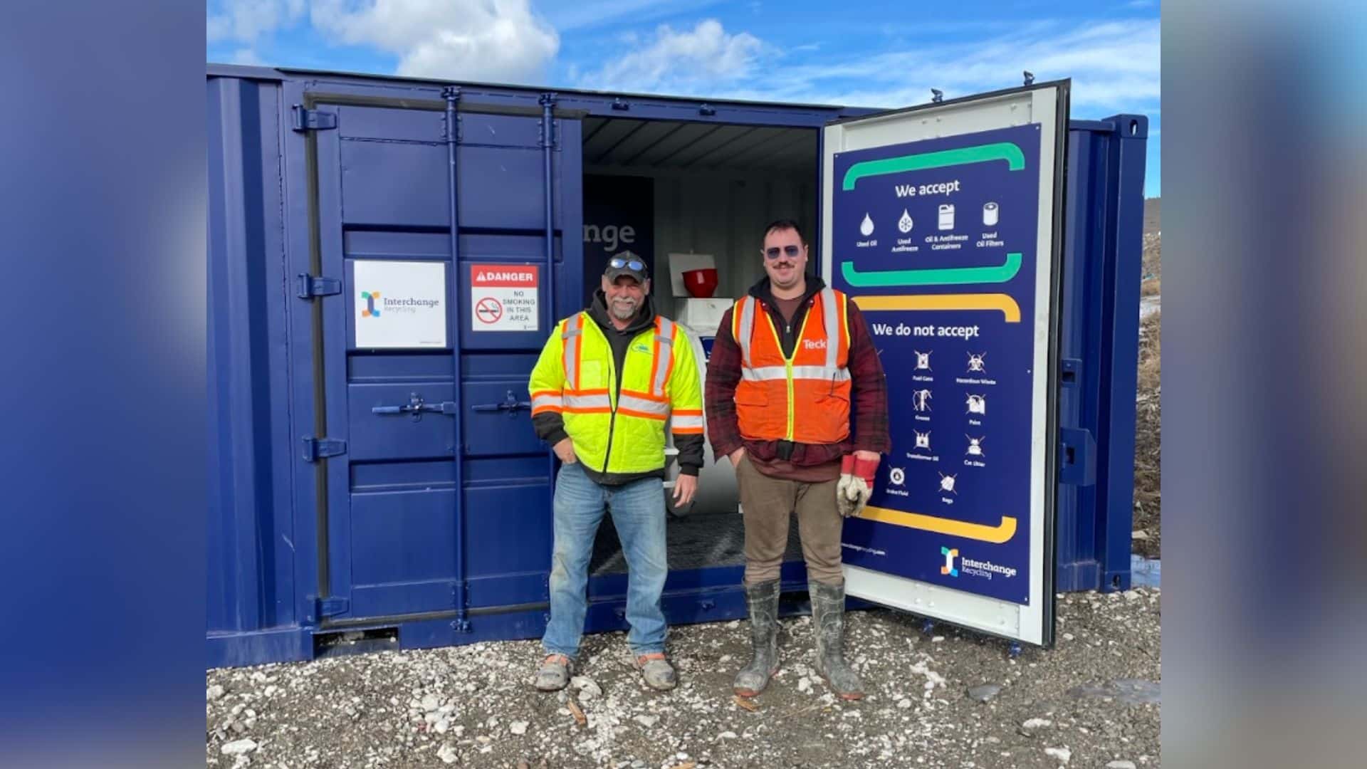 Two people standing outside of a large blue container for motor vehicle waste.