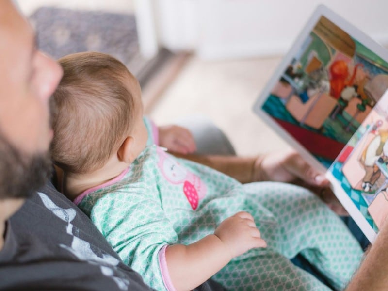 A dad reading a book to his young baby. (Picsea/Unsplash)